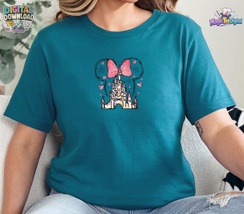 Pu&ograve; includere: Una t-shirt color verde acqua a maniche corte con una grafica centrale che raffigura la sagoma di un castello sormontata da un fiocco rosa a pois. Il design include contorni neri abbozzati di orecchie di topo, piccoli cuori rosa e stelle.