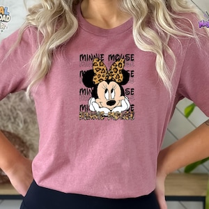 Pode incluir: Uma t-shirt malva de manga curta com um gr&aacute;fico de um rato de desenho animado com um la&ccedil;o de estampado de leopardo. O texto Minnie Mouse &eacute; repetido no fundo e impresso num padr&atilde;o de leopardo na parte inferior do design gr&aacute;fico.