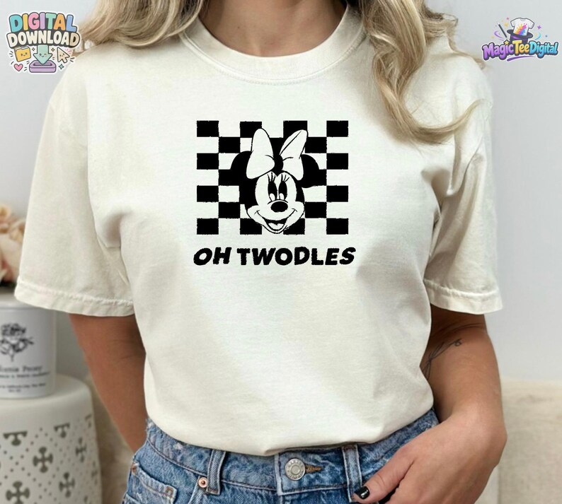 May include: A cream-coloured short-sleeve t-shirt featuring a black graphic of a cartoon mouse head with a bow centred over a black and white checkered square pattern. The text OH TWODLES is printed in black capital letters below the graphic.