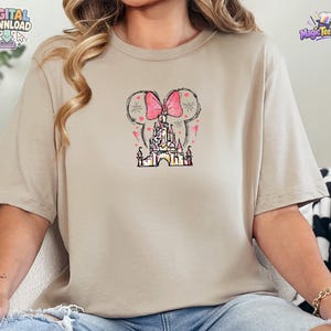 Pu&ograve; includere: Una t-shirt beige a maniche corte con una stampa grafica centrale raffigurante la sagoma di un castello sormontata da un grande fiocco rosa a pois e orecchie da topo. Il design include piccoli cuori rosa sparsi e sottili linee nere.