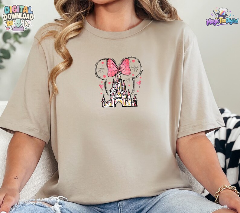 Pu&ograve; includere: Una t-shirt beige a maniche corte con una stampa grafica centrale raffigurante la sagoma di un castello sormontata da un grande fiocco rosa a pois e orecchie da topo. Il design include piccoli cuori rosa sparsi e sottili linee nere.