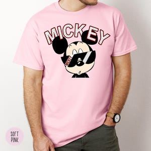 May include: A light pink t-shirt featuring a cartoon Mickey Mouse wearing sunglasses. The word "MICKEY" is printed above the character in a red and white font. The shirt is a solid color and has short sleeves.