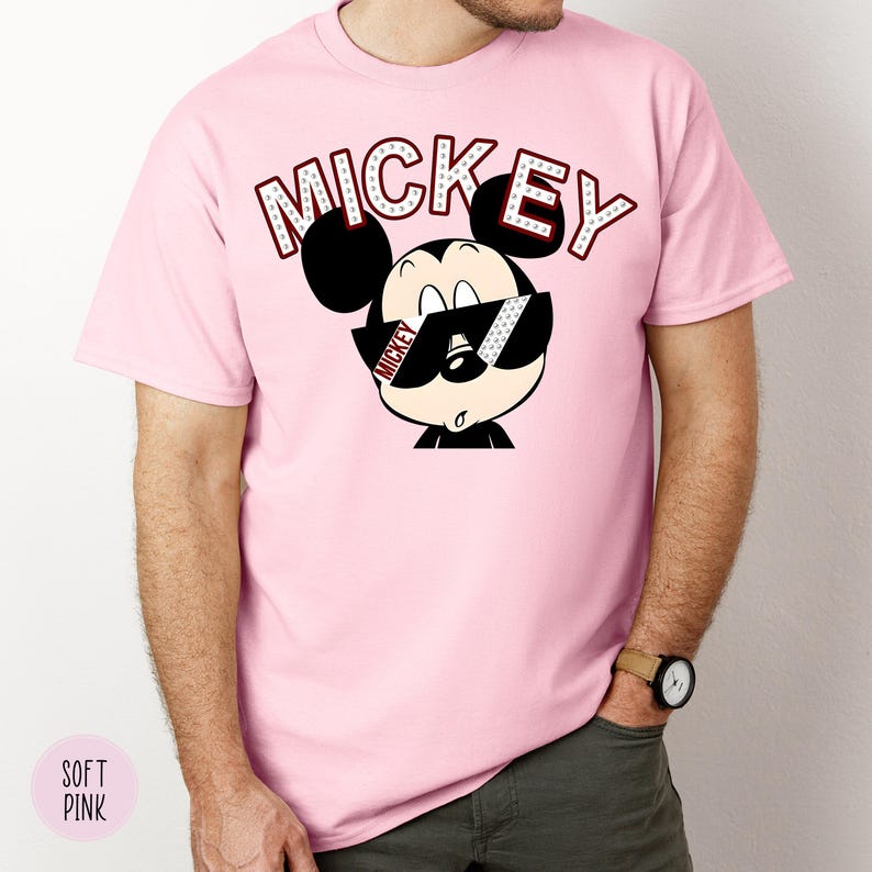 May include: A light pink t-shirt featuring a cartoon Mickey Mouse wearing sunglasses. The word "MICKEY" is printed above the character in a red and white font. The shirt is a solid color and has short sleeves.