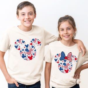 May include: Two children wearing cream-colored t-shirts. Each shirt features a Mickey Mouse head design made of red, white, and blue shapes. One shirt has a bow design. The shirts are perfect for patriotic celebrations.