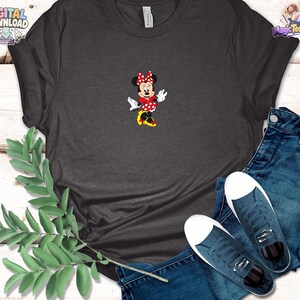Peut inclure: Un t-shirt gris fonc&eacute; &agrave; manches courtes avec un motif central repr&eacute;sentant une souris de dessin anim&eacute; portant une robe &agrave; pois rouges et blancs, un n&oelig;ud assorti et des chaussures jaunes. Accompagn&eacute; d'un jean bleu et de baskets en toile bleu fonc&eacute;.