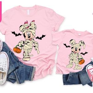 May include: Pink t-shirts featuring a mummy-themed Minnie Mouse and Mickey Mouse design, holding jack-o'-lanterns, with black bat silhouettes. The shirts are paired with blue jeans and white sneakers. The text "Ship Next Day" is visible.
