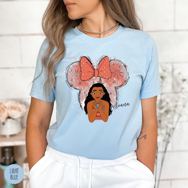 May include: Light blue t-shirt featuring a cartoon illustration of Moana. The design includes a large bow, Mickey Mouse ears, and the word "Moana" in script. The shirt is a short-sleeved crew neck.