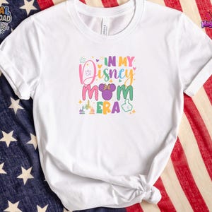 May include: A white short-sleeved t-shirt featuring a colorful graphic print that reads IN MY DISNEY MOM ERA. The design includes a purple mouse ear icon with a bow, a castle silhouette, and a hand gesture icon in various pastel colors.