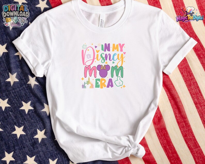 May include: A white short-sleeved t-shirt featuring a colorful graphic print that reads IN MY DISNEY MOM ERA. The design includes a purple mouse ear icon with a bow, a castle silhouette, and a hand gesture icon in various pastel colors.
