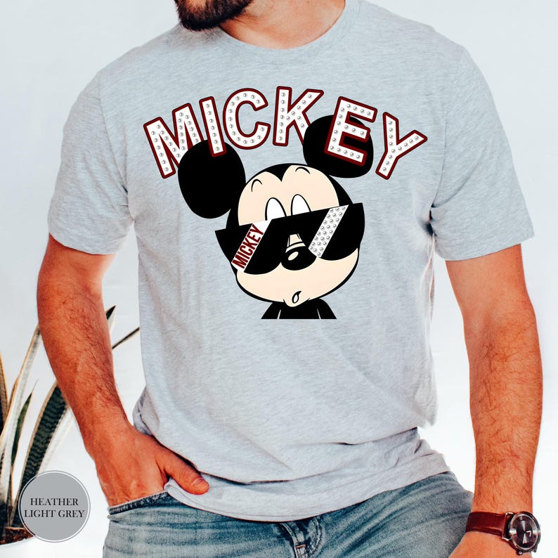 May include: Heather light grey t-shirt featuring a cartoon Mickey Mouse wearing sunglasses. The word "MICKEY" is printed above the character in a red and white font. The shirt is a short-sleeved crew neck.