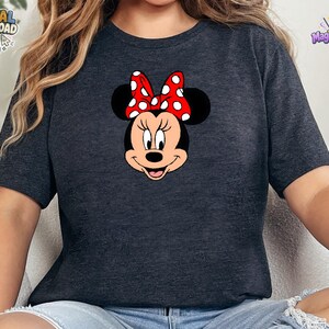 以下が含まれることがあります： ダークヘザーグレーの半袖Tシャツで、ミニーマウスの顔のグラフィックが描かれています。キャラクターは黒い耳、ピーチ色の顔をしており、頭の上部中央には白い水玉模様の鮮やかな赤いリボンが付いています。