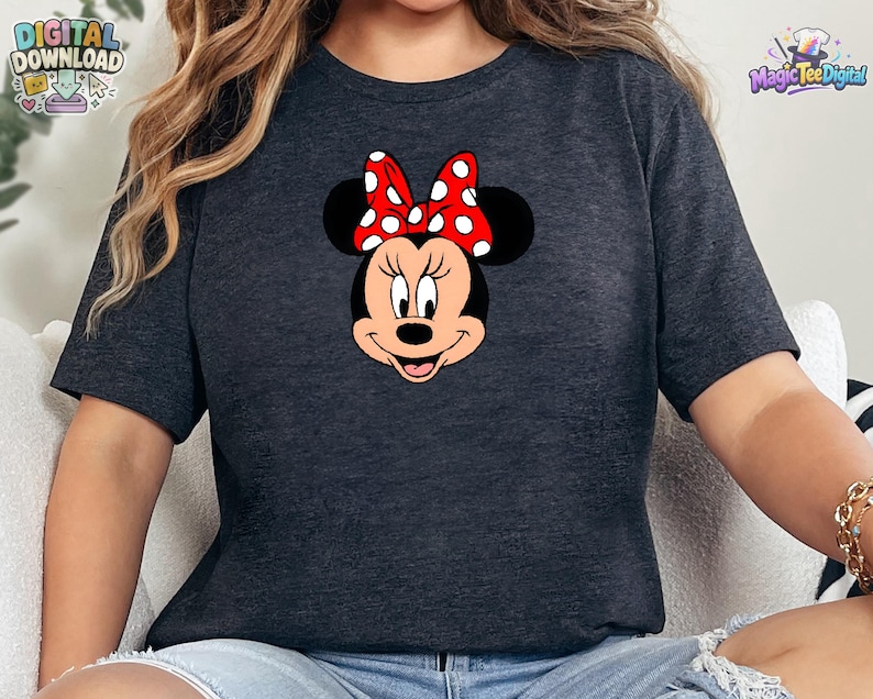 以下が含まれることがあります： ダークヘザーグレーの半袖Tシャツで、ミニーマウスの顔のグラフィックが描かれています。キャラクターは黒い耳、ピーチ色の顔をしており、頭の上部中央には白い水玉模様の鮮やかな赤いリボンが付いています。