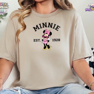 May include: A beige short-sleeve t-shirt featuring a graphic of Minnie Mouse wearing pink sunglasses and a pink polka dot dress. The text MINNIE is arched above the character, with EST. 1928 printed below, all in a black collegiate-style font.
