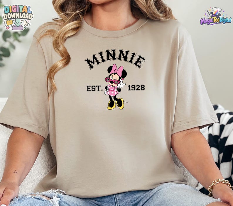 May include: A beige short-sleeve t-shirt featuring a graphic of Minnie Mouse wearing pink sunglasses and a pink polka dot dress. The text MINNIE is arched above the character, with EST. 1928 printed below, all in a black collegiate-style font.