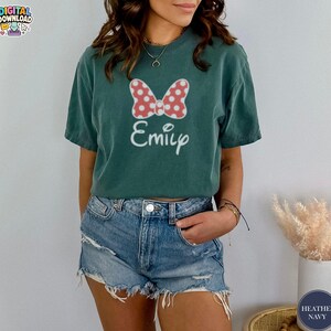 May include: A teal t-shirt featuring a red and white polka dot bow design and the name "Emily" in white script. The shirt is a short-sleeved, casual style.