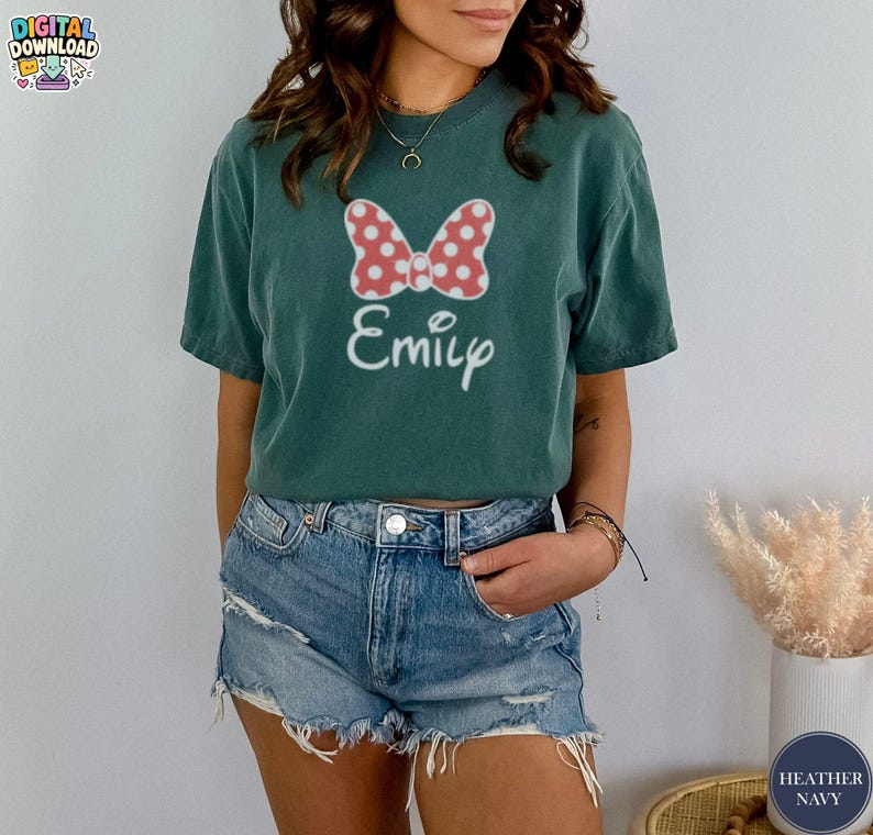 May include: A teal t-shirt featuring a red and white polka dot bow design and the name "Emily" in white script. The shirt is a short-sleeved, casual style.