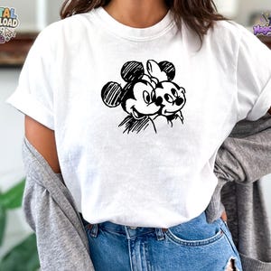 May include: A white short-sleeved t-shirt featuring a black ink-style sketch graphic of two cartoon mice characters positioned side-by-side on the chest. The design uses hatched lines to create the faces and round ears of the characters.
