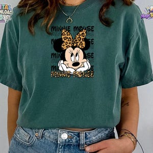 Pode incluir: Uma t-shirt de manga curta verde-azulado escuro com um gr&aacute;fico da Minnie Mouse com um la&ccedil;o de estampa de leopardo. O design inclui o texto "MINNIE MOUSE" repetido no fundo e impresso em estampa de leopardo na parte inferior.