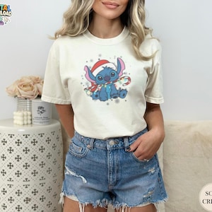 May include: Cream-colored t-shirt featuring a blue cartoon character wearing a Santa hat and scarf, holding a candy cane. The graphic includes snowflakes and festive lights. The shirt is paired with denim shorts.