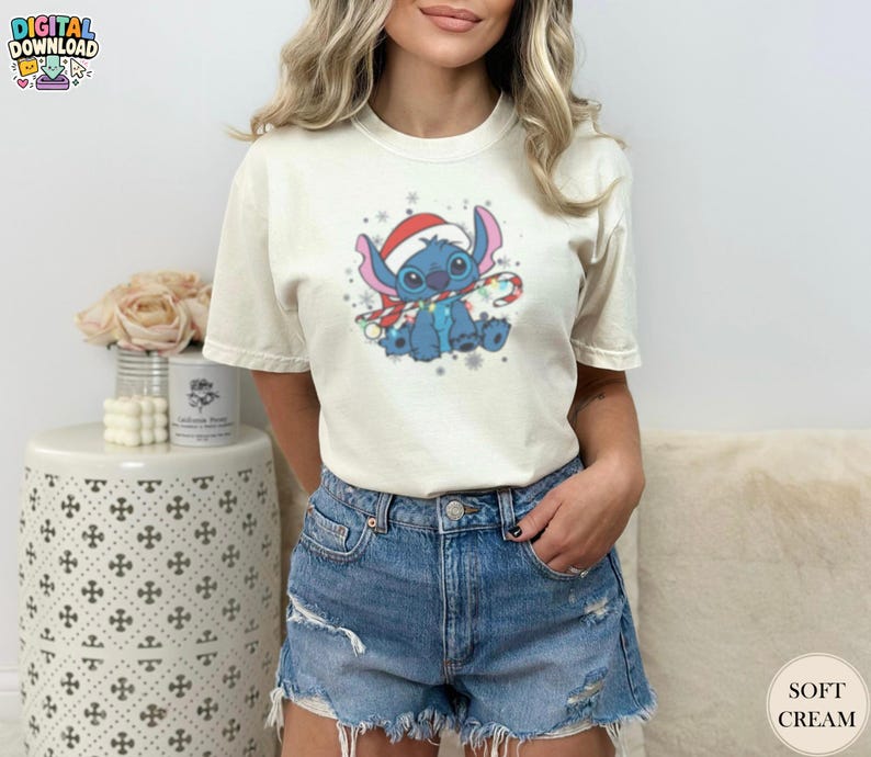 May include: Cream-colored t-shirt featuring a blue cartoon character wearing a Santa hat and scarf, holding a candy cane. The graphic includes snowflakes and festive lights. The shirt is paired with denim shorts.