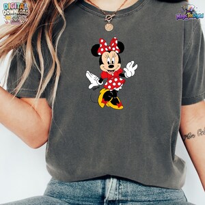 Peut inclure: Un t-shirt gris anthracite &agrave; manches courtes avec une impression graphique de Minnie Mouse portant une robe rouge &agrave; pois blancs, un n&oelig;ud rouge assorti et des chaussures jaunes. Le personnage est plac&eacute; au centre du v&ecirc;tement.