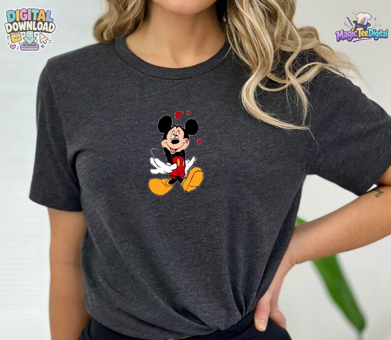 May include: A dark heather gray short-sleeve t-shirt featuring a centered graphic of Mickey Mouse. The character is depicted with a smiling expression, wearing red shorts and yellow shoes, surrounded by small red hearts floating above the head.