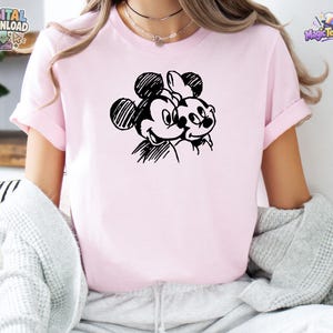 May include: A light pink short-sleeved t-shirt featuring a black line-art graphic of two cartoon mouse characters positioned side-by-side. The design uses a sketch-like style with visible hatching lines to define the faces and ears of the characters.