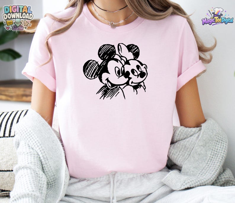 May include: A light pink short-sleeved t-shirt featuring a black line-art graphic of two cartoon mouse characters positioned side-by-side. The design uses a sketch-like style with visible hatching lines to define the faces and ears of the characters.