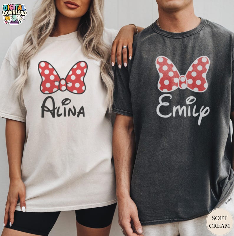 May include: Two matching t-shirts, one cream and one charcoal, featuring a red polka-dot bow design. The cream shirt has the name "Alina" in script, while the charcoal shirt says "Emily". Both shirts are soft cream colored.