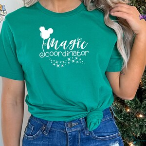 May include: A kelly green short-sleeve t-shirt featuring a white graphic print of a mouse-ear balloon and the text "magic coordinator" in a cursive font, accompanied by a trail of small sparkling stars below the lettering.