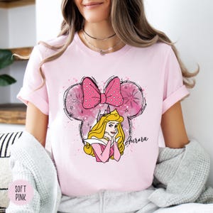 May include: A pale pink t-shirt with a cartoon illustration of Princess Aurora. The design includes a pink polka-dot bow, a castle, and the name "Aurora" in cursive. The shirt has short sleeves.