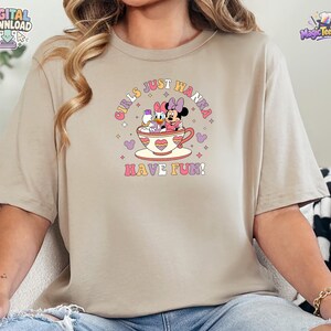 May include: A beige short-sleeve t-shirt featuring a graphic of Minnie Mouse and Daisy Duck sitting in a teacup. The text "GIRLS JUST WANNA HAVE FUN!" is printed in a retro font above and below the characters, surrounded by small stars and hearts.