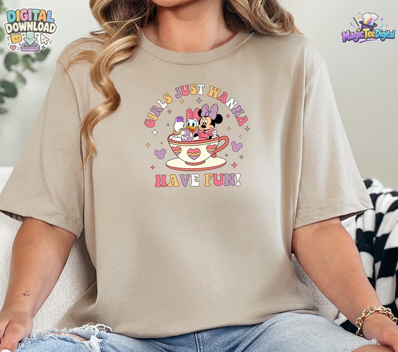 May include: A beige short-sleeve t-shirt featuring a graphic of Minnie Mouse and Daisy Duck sitting in a teacup. The text "GIRLS JUST WANNA HAVE FUN!" is printed in a retro font above and below the characters, surrounded by small stars and hearts.