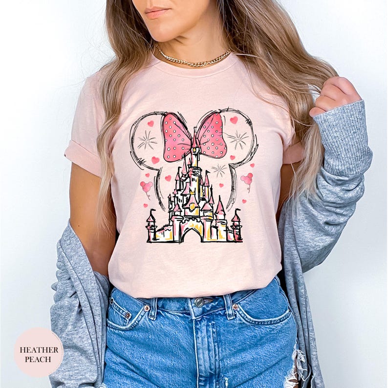 Puede incluir: Camiseta melocot&oacute;n claro con un dise&ntilde;o de castillo caprichoso, un lazo rosa y orejas de Mickey Mouse. El dise&ntilde;o incluye corazones rosas y una ilustraci&oacute;n de castillo. El texto "HEATHER PEACH" est&aacute; en la parte inferior izquierda.