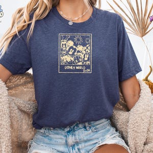 K&ouml;nnte beinhalten: Ein kurz&auml;rmeliges T-Shirt in meliertem Marineblau mit einem cremefarbenen Grafikdruck eines Polaroid-Rahmens, der Disney-Figuren und Feuerwerk zeigt, mit dem deutlich lesbaren Text "DISNEY WORLD 2024" am unteren Rand des Rahmens.