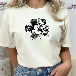 May include: A cream-colored short-sleeve t-shirt featuring a black ink-style sketch graphic of two cartoon mice heads positioned side-by-side on the chest. The design uses hatched lines to create the character features and outlines.