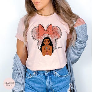 May include: Heather peach-colored t-shirt featuring a cartoon illustration of Moana within a Mickey Mouse ear outline. The design includes a coral-colored bow with white polka dots and the name "Moana" in script. The shirt is made of soft material.