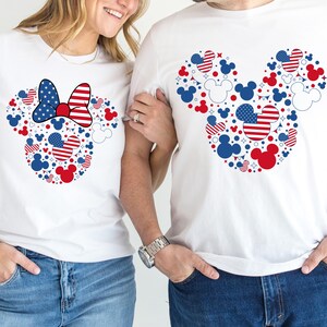 May include: White t-shirts featuring a Mickey Mouse head design composed of red, white, and blue shapes and patterns. One shirt has a Minnie Mouse bow. The shirts are worn with blue jeans.