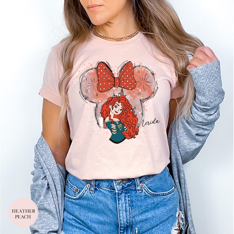 Puede incluir: Camiseta melocot&oacute;n claro con una ilustraci&oacute;n de Merida de Brave. El dise&ntilde;o incluye un lazo rojo con lunares blancos, orejas de Mickey Mouse y el nombre "Merida". La camiseta es de color rosa claro.