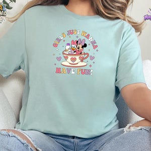May include: A light mint green short-sleeve t-shirt featuring a graphic of Minnie Mouse and Daisy Duck sitting in a teacup. The text "GIRLS JUST WANNA HAVE FUN!" is printed in a retro font around the characters, accented by small stars and hearts.