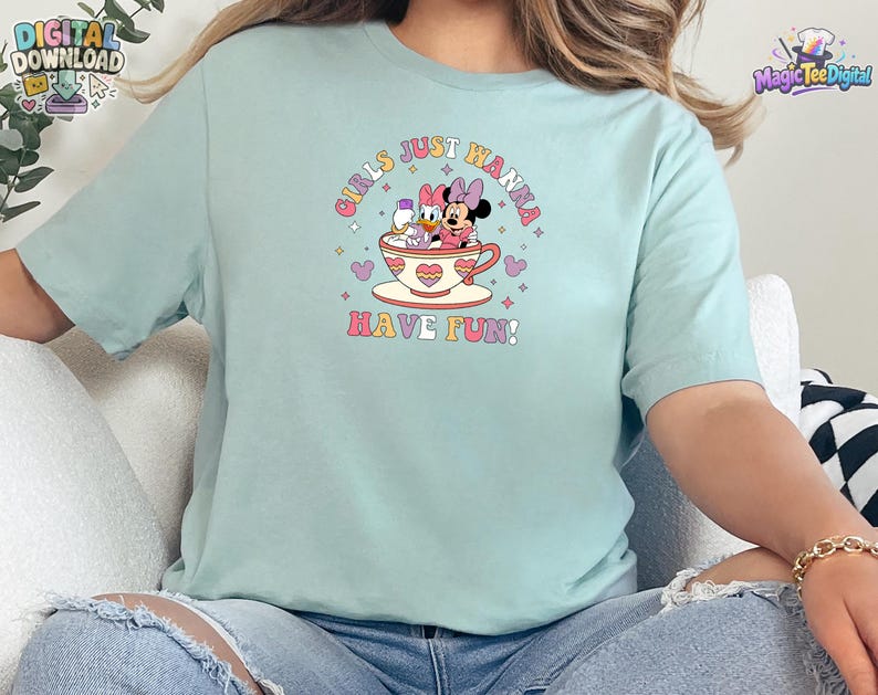 May include: A light mint green short-sleeve t-shirt featuring a graphic of Minnie Mouse and Daisy Duck sitting in a teacup. The text "GIRLS JUST WANNA HAVE FUN!" is printed in a retro font around the characters, accented by small stars and hearts.