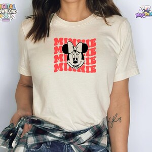 May include: A cream-colored short-sleeve t-shirt featuring a graphic print of Minnie Mouse centered on the chest. The design includes the name Minnie repeated in a red, wavy, stacked font behind the black and white character illustration.