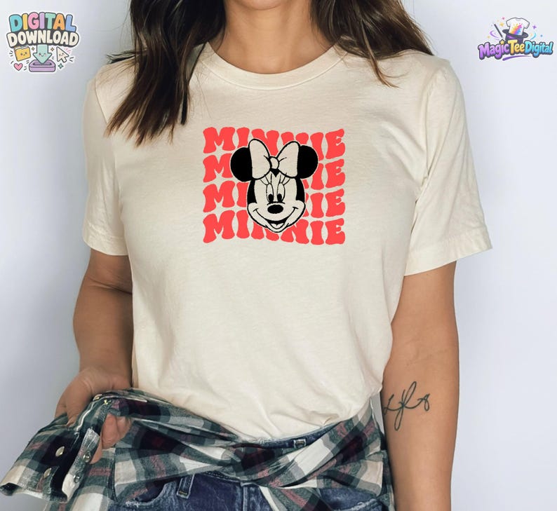 May include: A cream-colored short-sleeve t-shirt featuring a graphic print of Minnie Mouse centered on the chest. The design includes the name Minnie repeated in a red, wavy, stacked font behind the black and white character illustration.