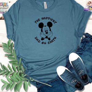 Puede incluir: Una camiseta azul pizarra con un gr&aacute;fico negro de un rat&oacute;n de dibujos animados con las manos en la cara, rodeado por el texto THE HAPPIEST DAD ON EARTH. La camiseta se combina con vaqueros azul oscuro y zapatillas de lona azules a juego.