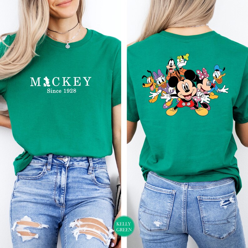 Puede incluir: Camiseta verde Kelly con el texto "Mickey Since 1928" en la parte delantera. La parte trasera de la camiseta muestra un gr&aacute;fico colorido de Mickey Mouse y sus amigos. La camiseta se combina con unos vaqueros azules.