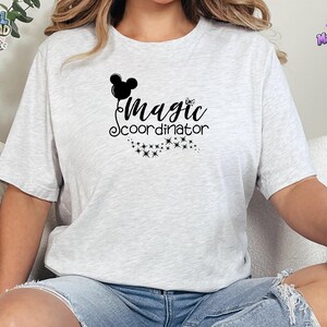 May include: A light gray heathered t-shirt featuring a black graphic print on the chest. The design includes a silhouette of a mouse-ear balloon, the text "Magic coordinator" in a cursive script, and a trail of small sparkling star icons.