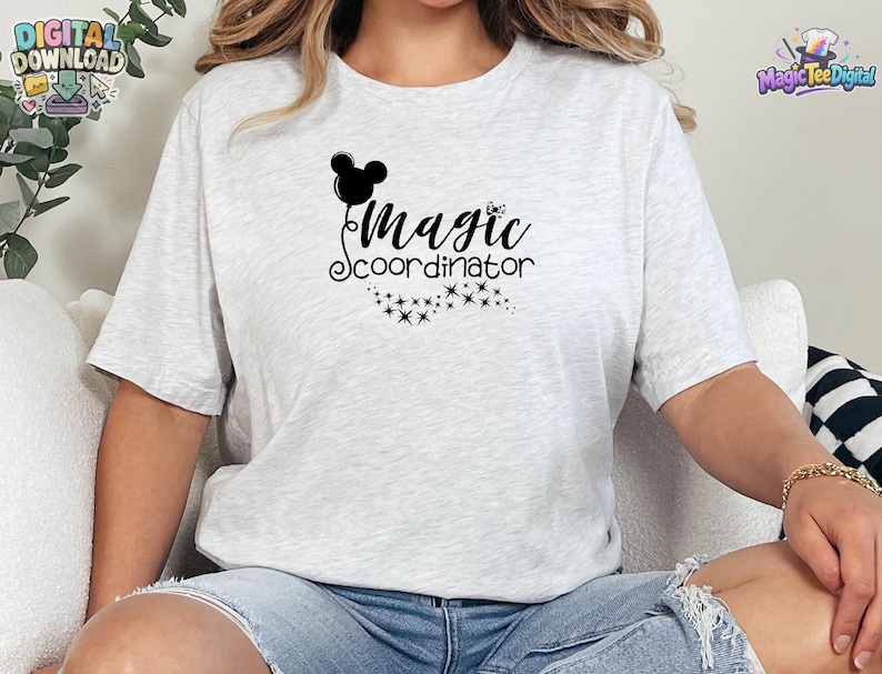May include: A light gray heathered t-shirt featuring a black graphic print on the chest. The design includes a silhouette of a mouse-ear balloon, the text "Magic coordinator" in a cursive script, and a trail of small sparkling star icons.