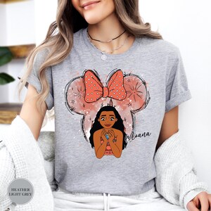 May include: Heather light grey t-shirt featuring a graphic of Moana with a Minnie Mouse-inspired design. The design includes a coral bow with white polka dots, a Moana character illustration, and the name "Moana" in script.
