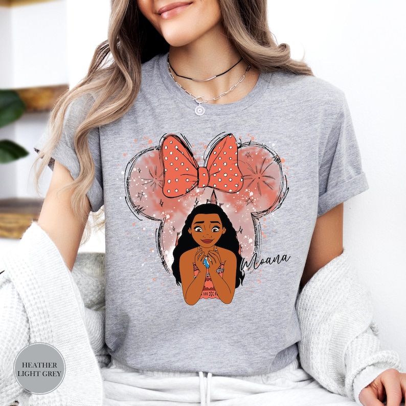 May include: Heather light grey t-shirt featuring a graphic of Moana with a Minnie Mouse-inspired design. The design includes a coral bow with white polka dots, a Moana character illustration, and the name "Moana" in script.