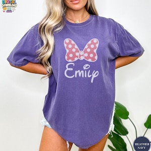 May include: A purple t-shirt featuring a pink polka-dot bow and the name "Emily" in white script. The shirt has short sleeves and a relaxed fit. The words "Digital Download" are in the top left corner.